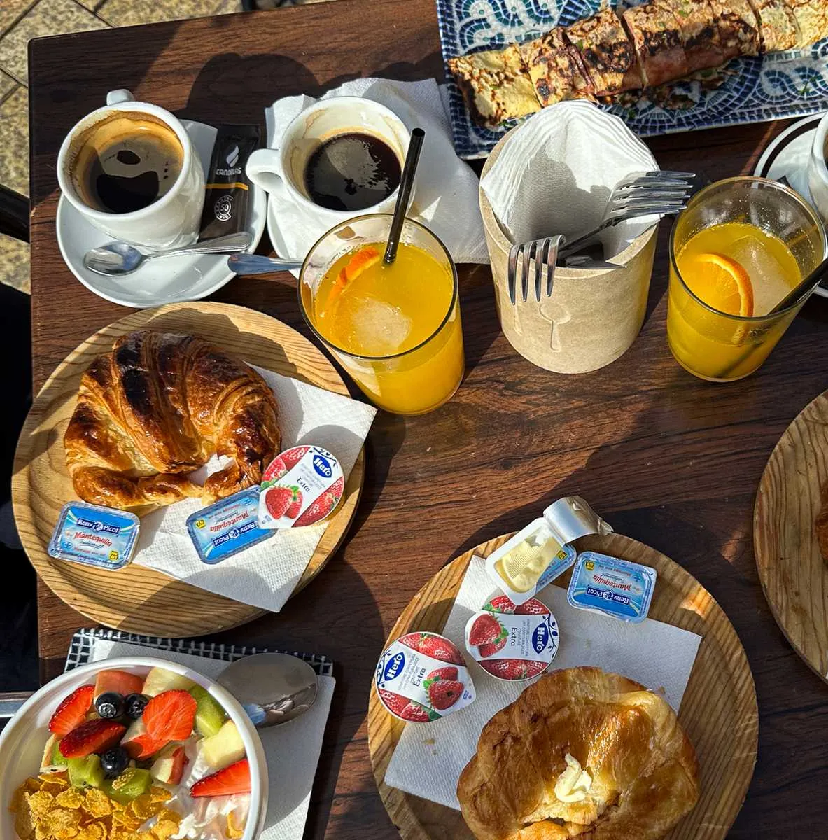A breakfast table in the sun: croissants with butter and jam, two coffees, orange juice and a bowl of yoghurt with fruit